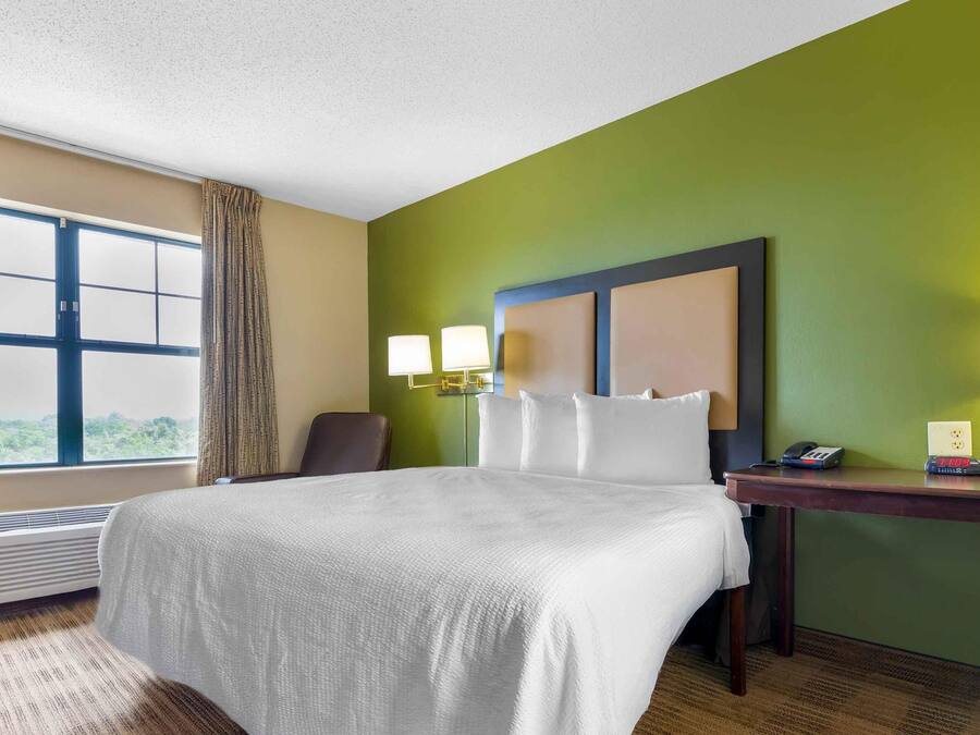 Quarto de hotel com uma cama grande, uma mesa com luminária, uma cadeira e uma janela com cortinas.