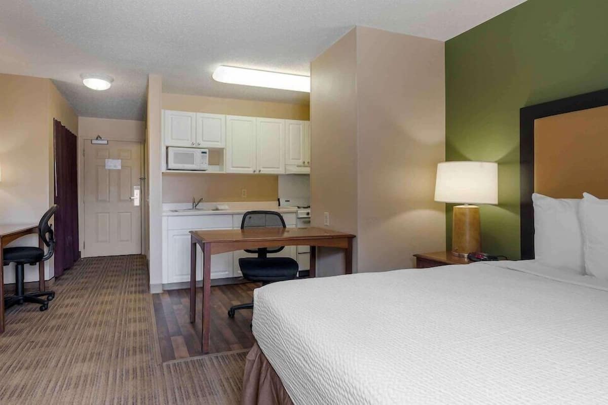 A hotel room with a bed, desk, chair, kitchenette, and a lamp.