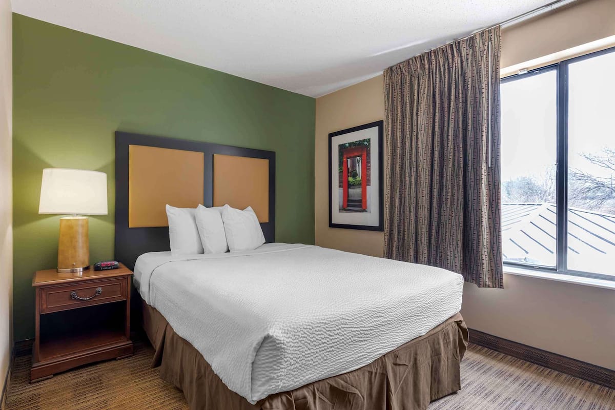 A hotel room with a bed, a nightstand with a lamp, a framed picture on the wall, and a window with curtains.