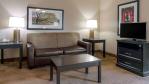 A hotel room with a brown leather sofa, a small coffee table, two bedside tables with lamps, a flat-screen TV on a black stand, and framed artwork on the walls. - Extended Stay America Select Suites Dallas Farmers Branch (Dallas)