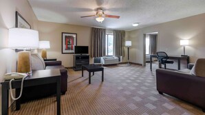 A hotel room with a ceiling fan, a desk, a chair, a sofa, a TV, a telephone, and a window with curtains. - Extended Stay America Select Suites Dallas Farmers Branch (Dallas)