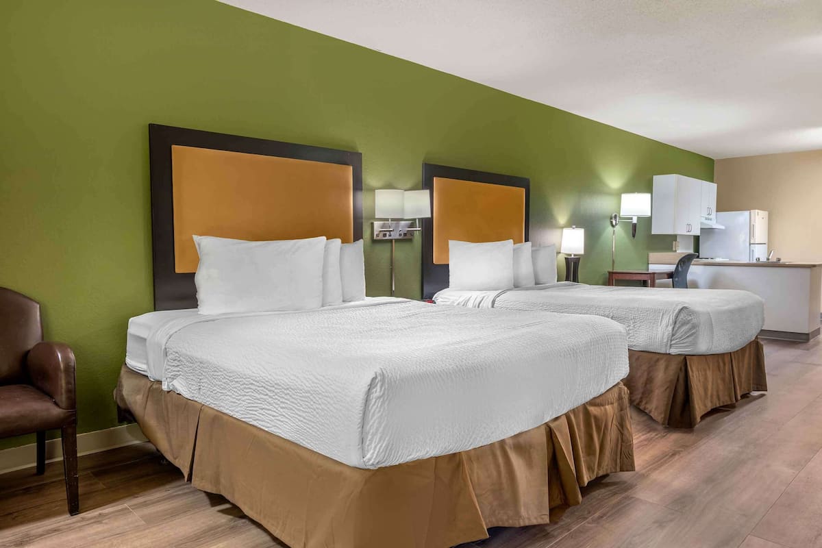 A hotel room with two beds, a desk, a chair, and a kitchenette.