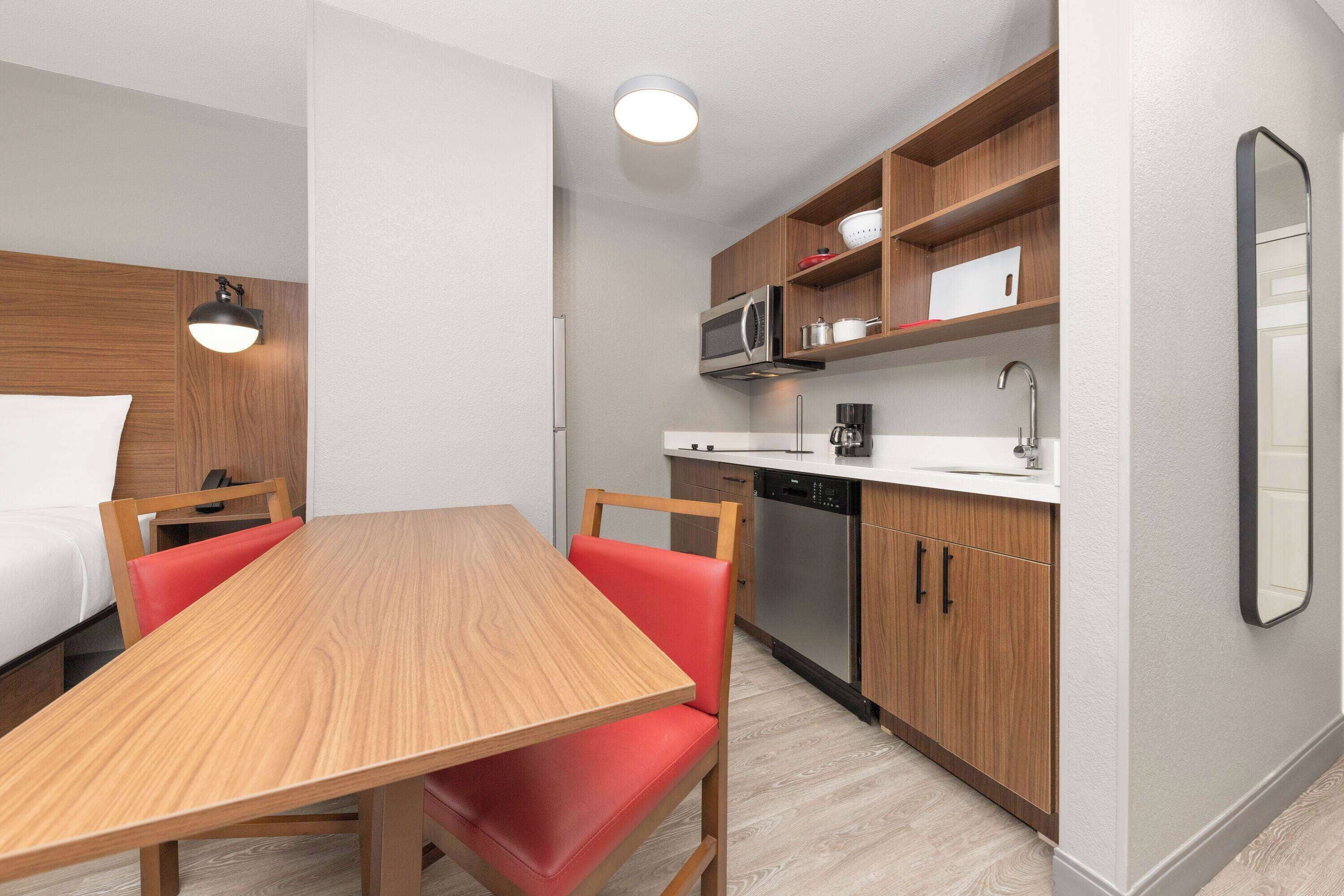 Une chambre d’hôtel compacte comprenant une table à manger en bois, des chaises rembourrées de couleur rouge, un four à micro-ondes, une machine à café et un lave-vaisselle.