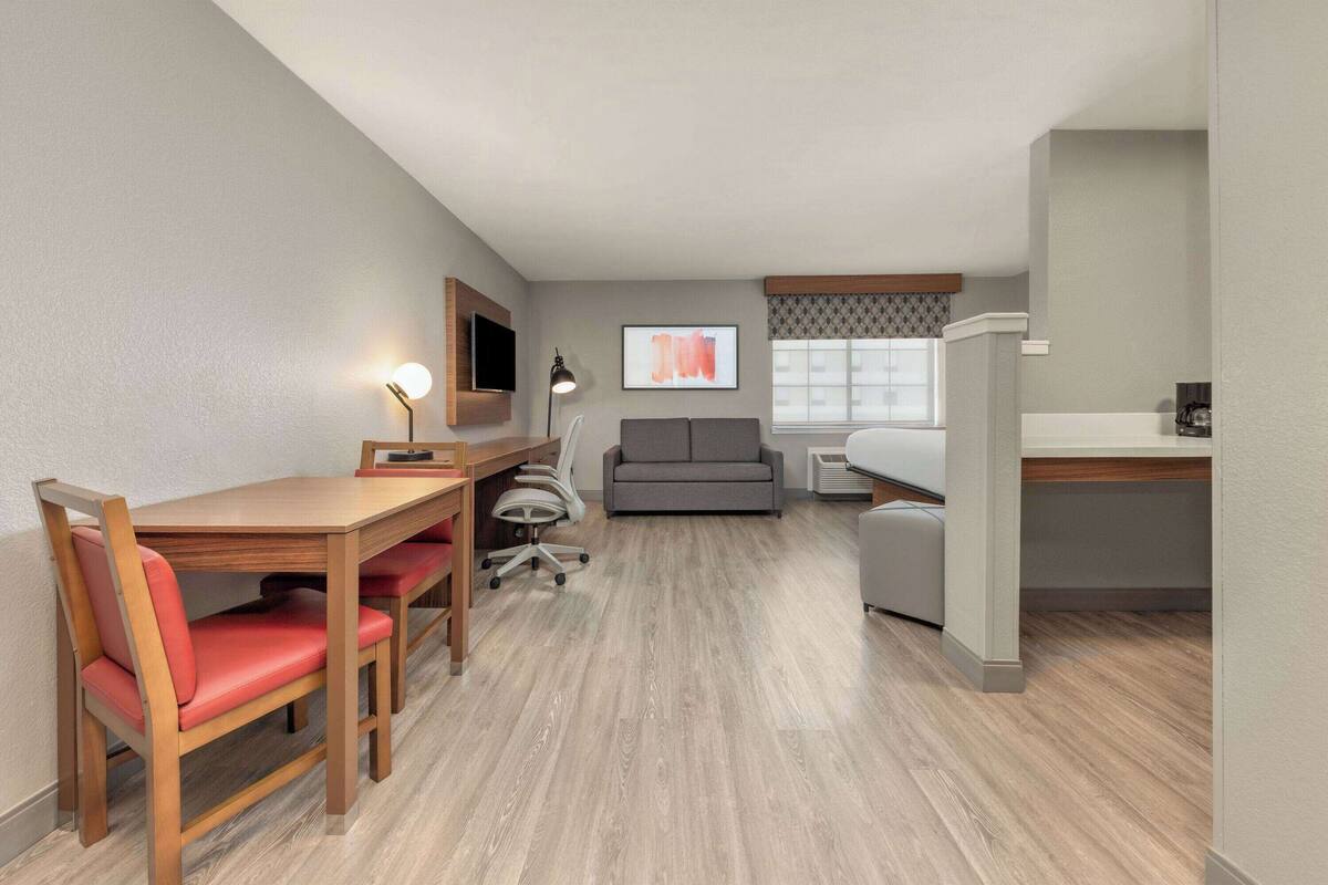 A compact hotel room with a wooden desk, a red chair, a gray sofa, and a small kitchenette.