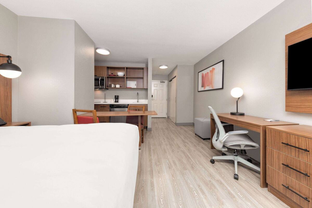 A hotel room with a bed, a desk, a chair, a TV, and a kitchenette.