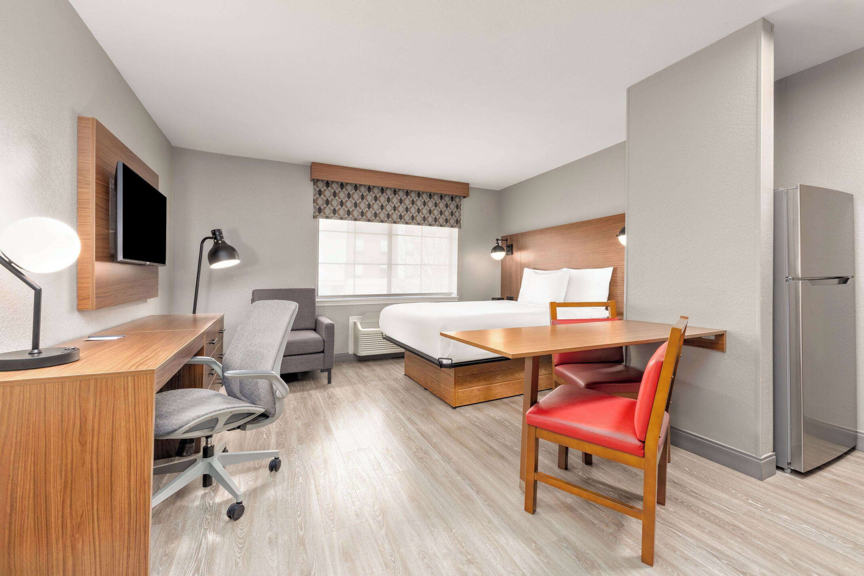 A hotel room with a bed, desk, chair, and a refrigerator.