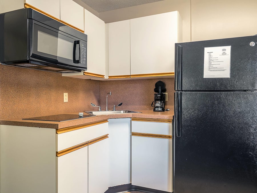 Una cocina compacta con armarios blancos, un refrigerador negro, un microondas, una cafetera y un lavamanos.