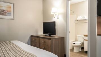 A hotel room with a bed, a TV on a wooden cabinet, a wall-mounted lamp, and a bathroom visible through an open door.