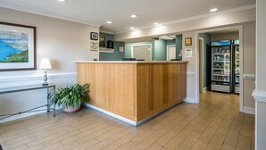 A reception area with a wooden reception desk, a potted plant, a framed painting, and a refrigerator stocked with beverages. - Suburban Studios of Wilmington (Wilmington)