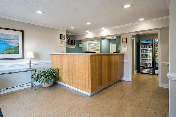 A reception area with a wooden reception desk, a potted plant, a framed painting, and a refrigerator stocked with beverages.