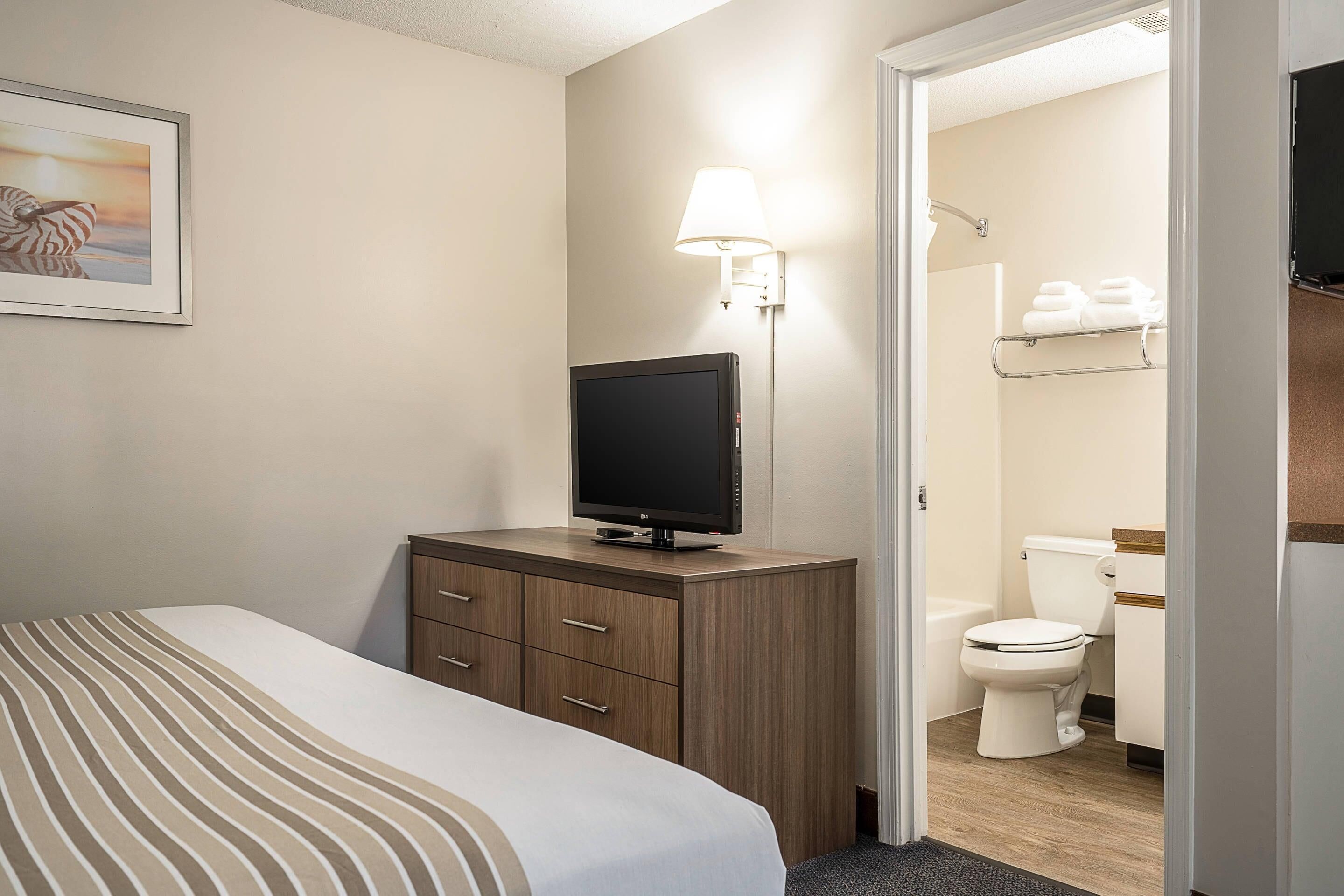 A hotel room with a bed, a TV on a wooden cabinet, a wall-mounted lamp, and a bathroom visible through an open door.