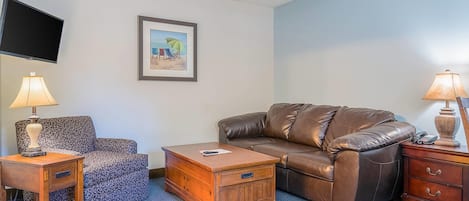 Una sala de estar con un sofá de cuero marrón, un sillón estampado, una mesa de centro de madera y un televisor montado en la pared.