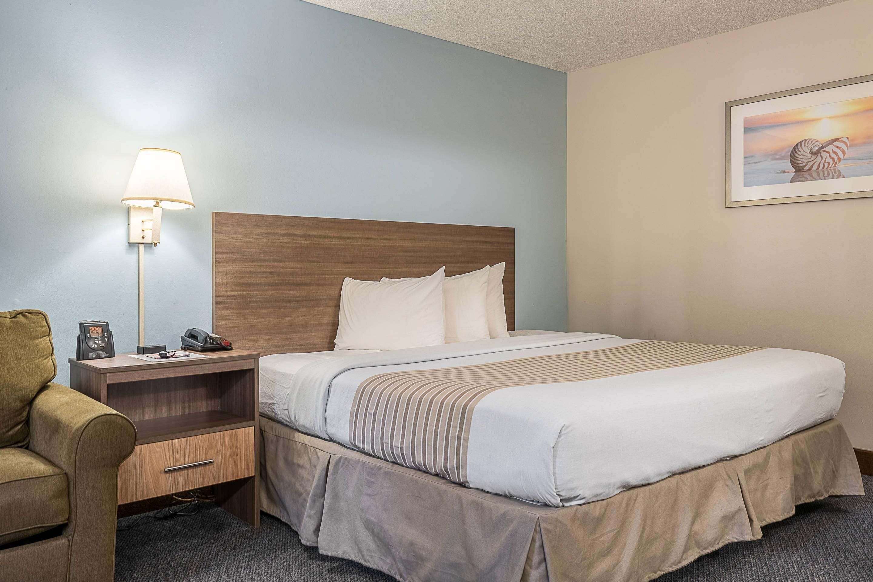 A hotel room with a bed, bedside table, lamp, chair, and a painting on the wall.