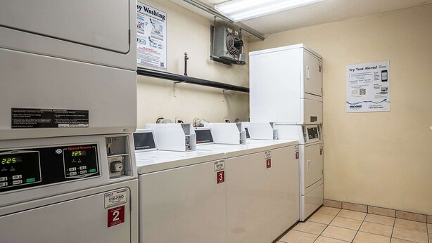A laundry room with multiple washing machines and dryers, numbered signs, and a poster advertising text alerts. - Suburban Studios of Wilmington (Wilmington)