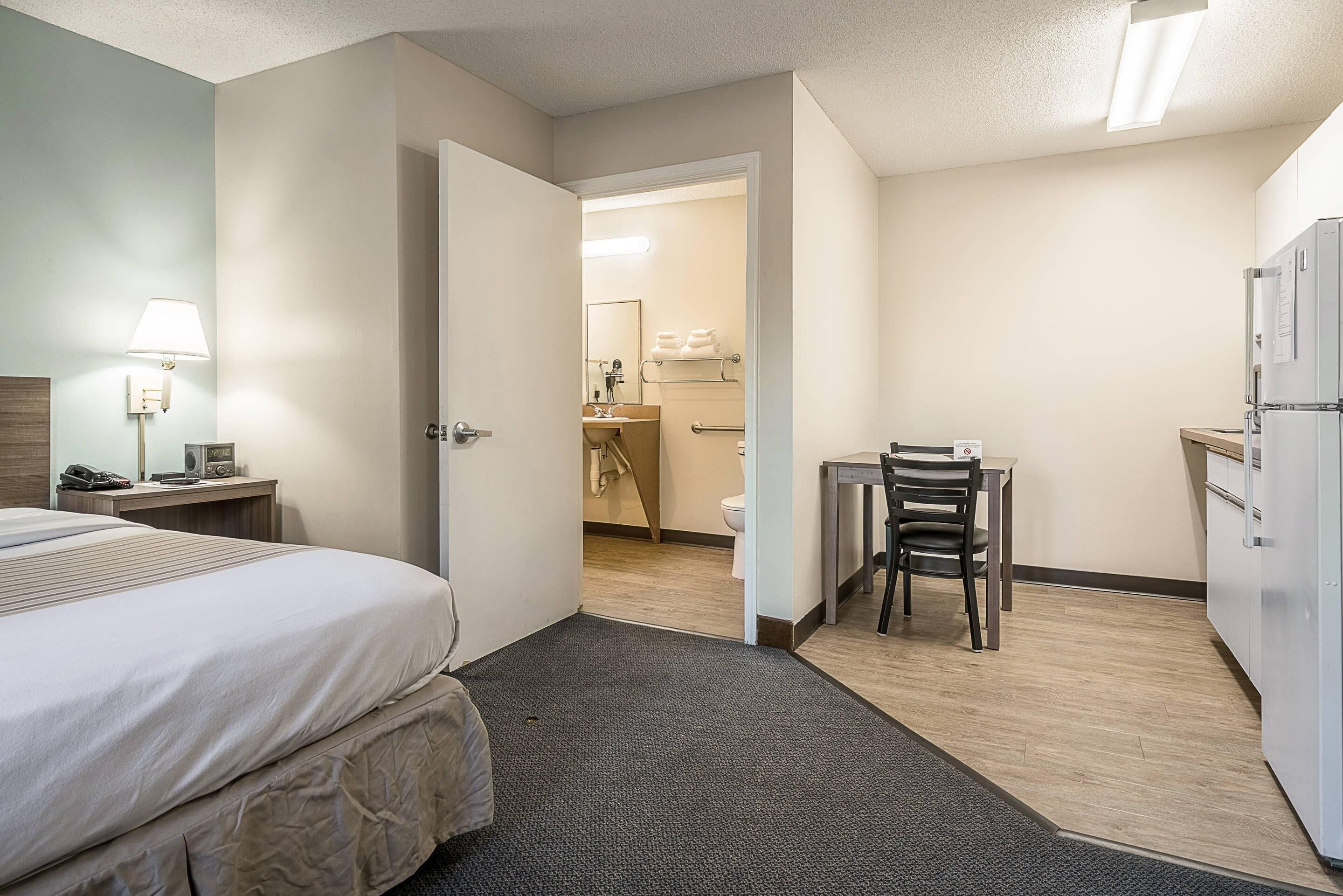 A hotel room with a bed, a desk, a chair, a refrigerator, and a bathroom visible through an open door.