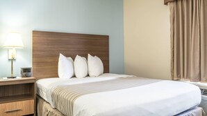 A hotel room with a wooden headboard, a bedside table with a lamp, a radio, and a bed with white bedding and striped pillows. - Suburban Studios of Wilmington (Wilmington)