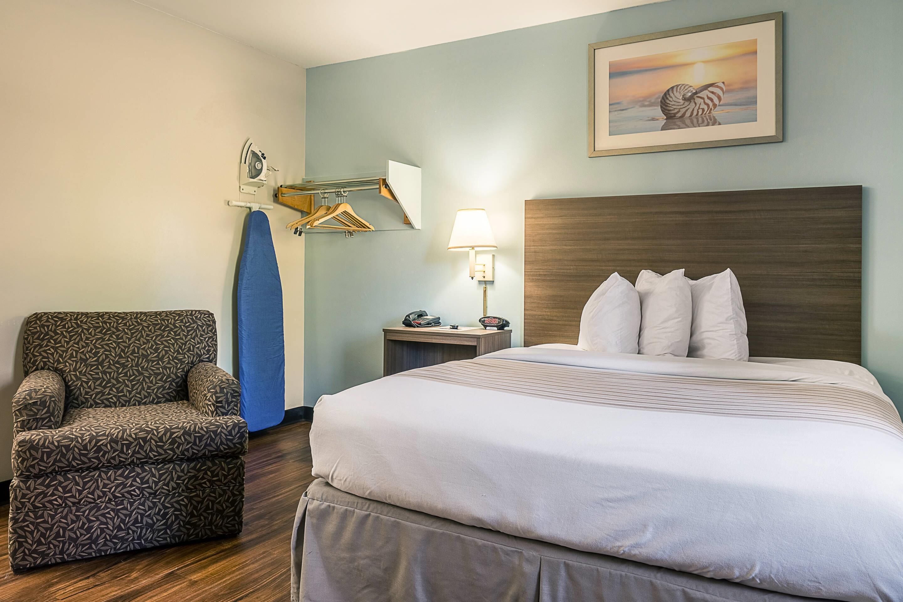 A hotel room with a bed, a chair, a desk with a lamp, and a painting on the wall.