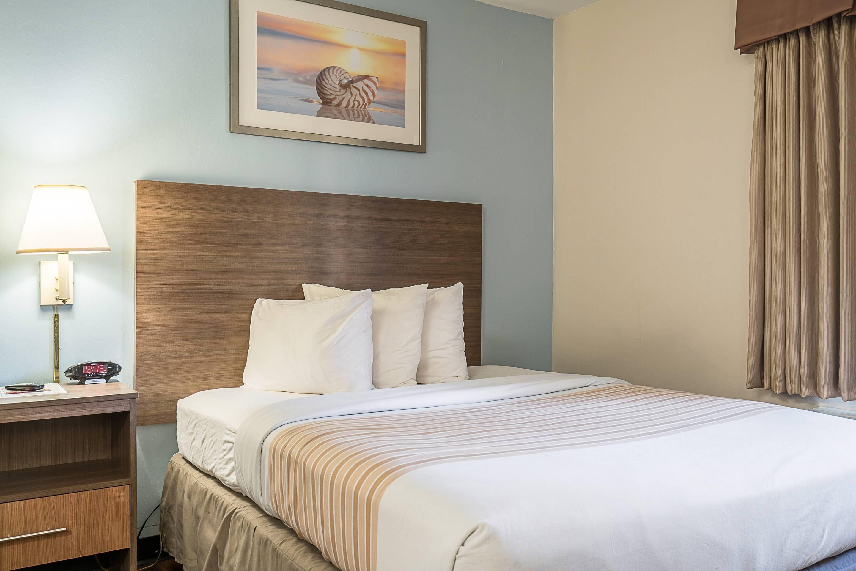 Una habitación de hotel con una cama, una mesita de noche con una lámpara, un reloj y un cuadro en la pared.
