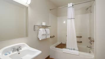 A bathroom with a white sink, a mirror, a shower with a white curtain, and a wooden shelf with towels.
