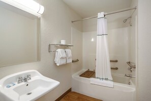 Un baño con lavamanos blanco, un espejo, una ducha con cortina blanca y un estante de madera con toallas.