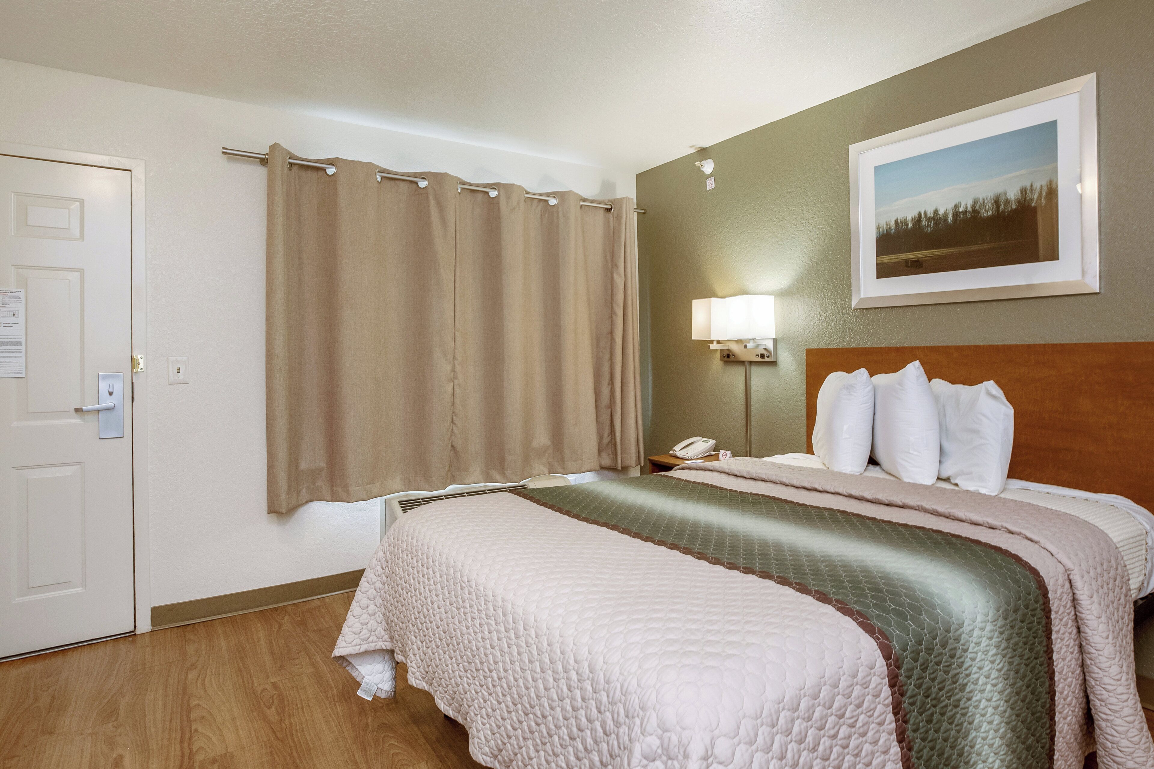 Quarto de hotel com uma cama, mesa de cabeceira, telefone, abajur, porta e um quadro pendurado na parede.