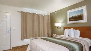 Habitación de hotel con cama, mesita de noche, teléfono, lámpara, puerta y un cuadro colgado en la pared.
