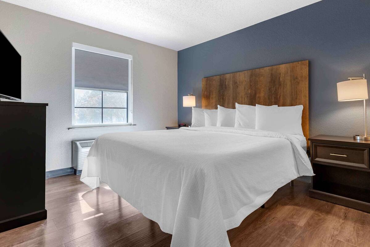 Una cama bien tendida con sábanas blancas, un cabecero de madera, una lámpara de noche y una mesita de noche con cajón.