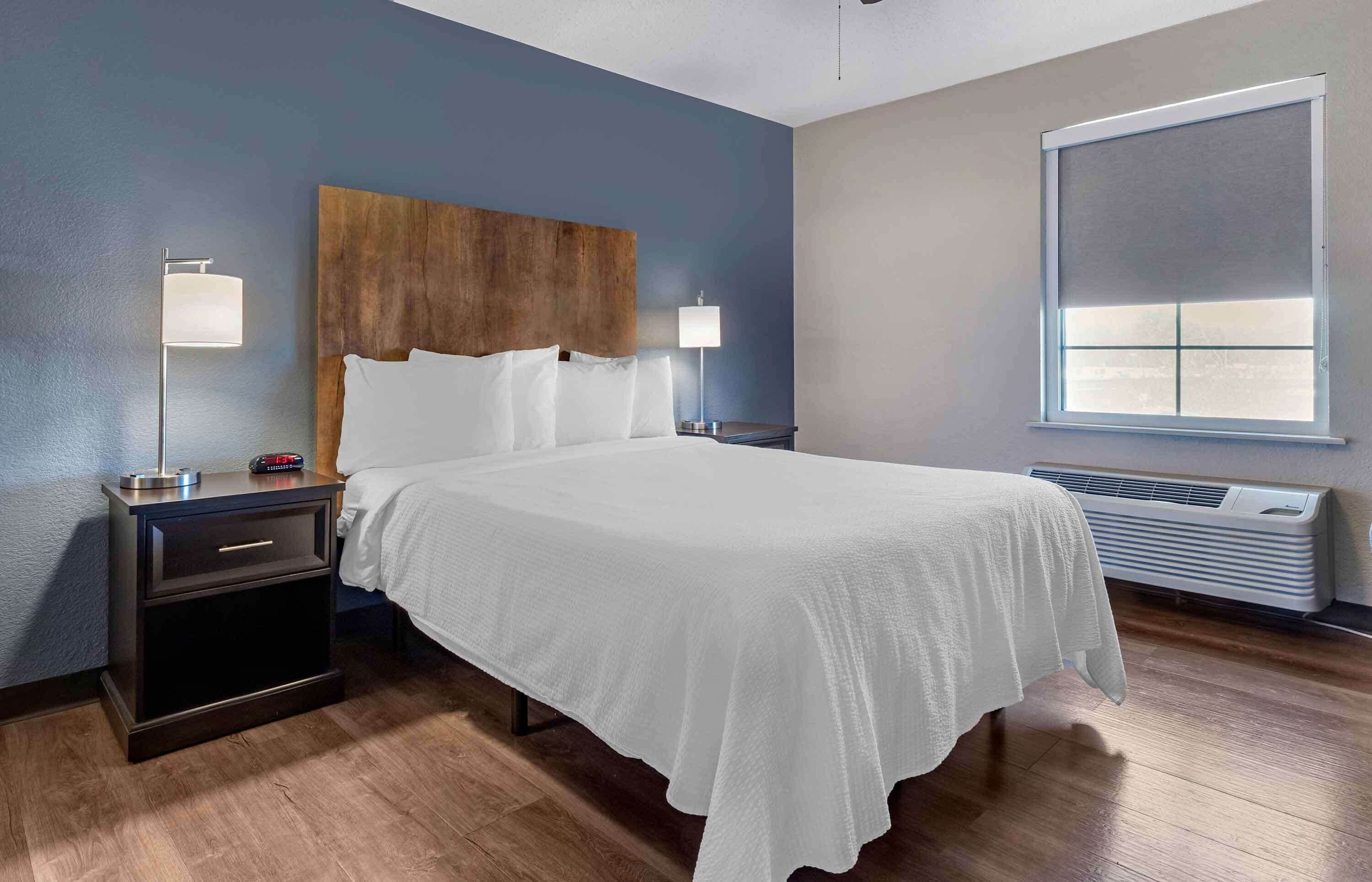 Quarto de hotel com uma cama grande, duas mesas de cabeceira com luminárias, uma cabeceira de madeira, uma janela com persianas e um ar-condicionado fixado na parede.