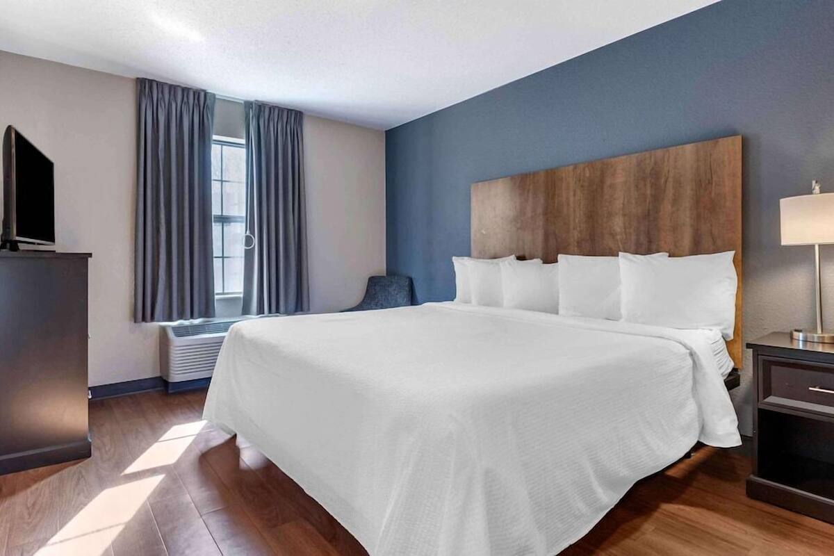Habitación de hotel con una cama grande, un televisor sobre un mueble bajo, un escritorio con lámpara y una ventana con cortinas.
