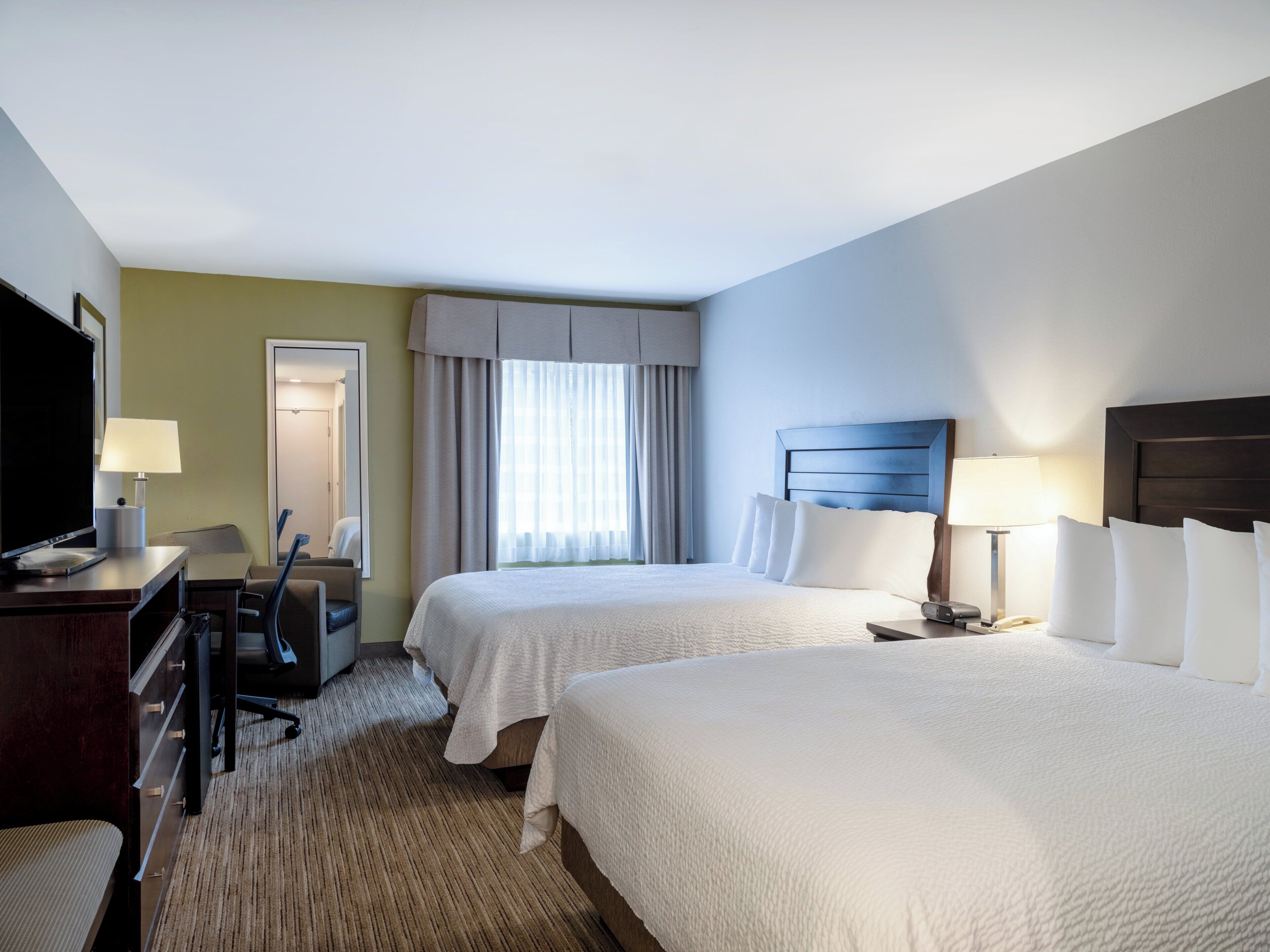 A hotel room with two beds, a desk, a chair, a TV, and a window with curtains.