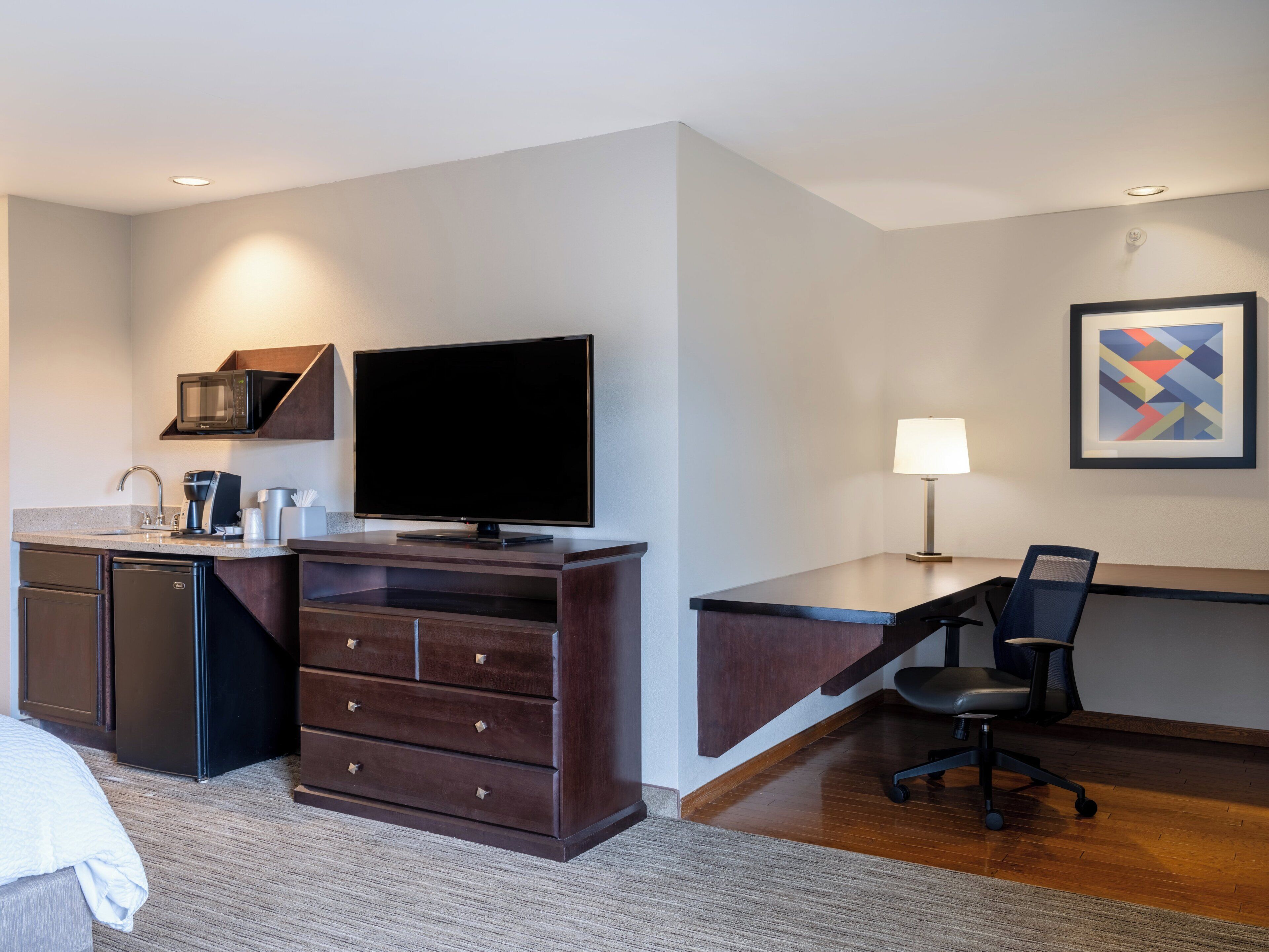 A hotel room with a bed, a desk with a chair, a television, a microwave, a coffee maker, and a small cabinet.