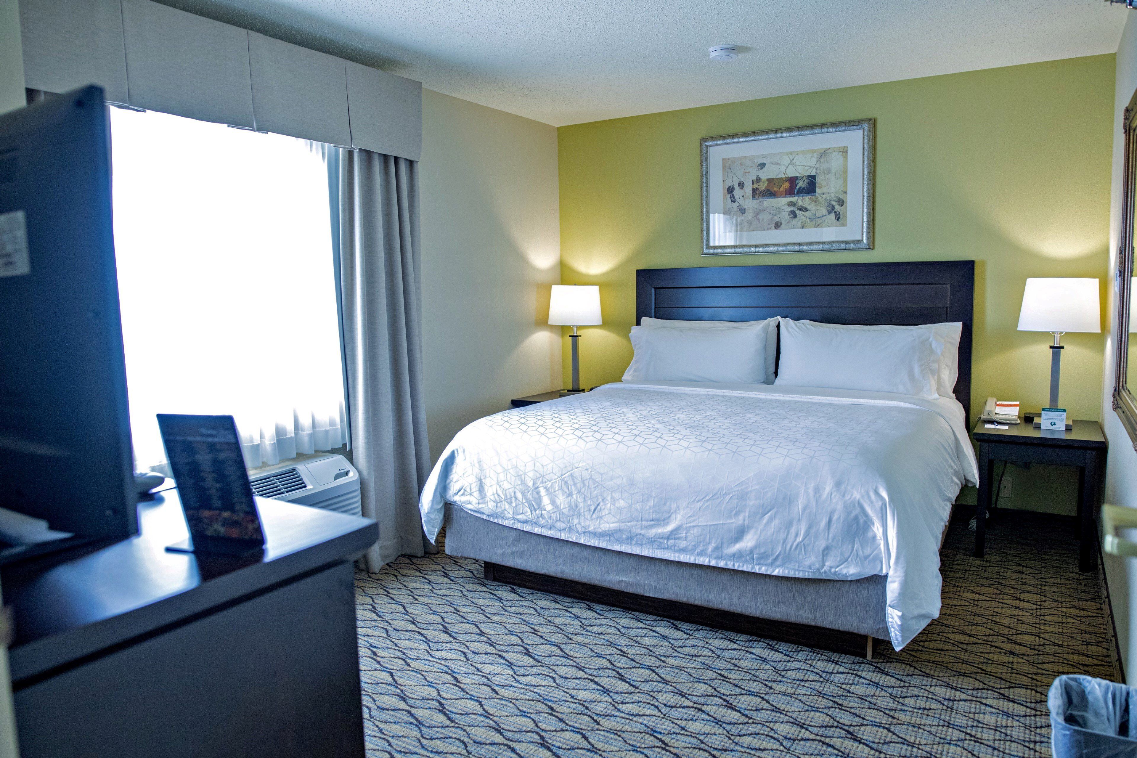 A hotel room with a large bed, two bedside tables, a desk with a computer, and a window with curtains.