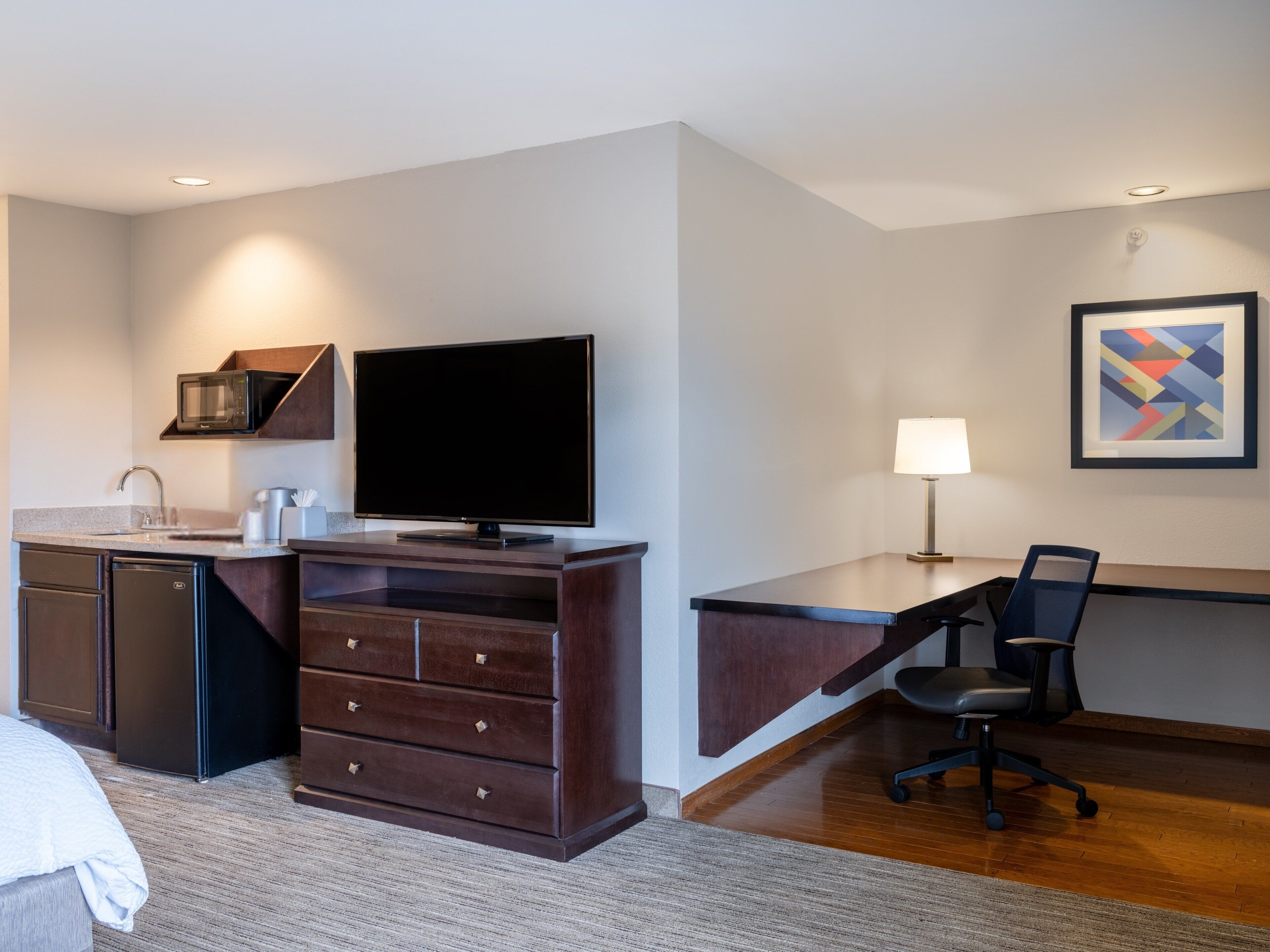 A hotel room with a kitchenette, a flat-screen TV, a desk with a chair, a lamp, and a framed abstract art piece on the wall.