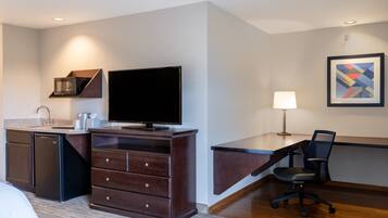 A hotel room with a kitchenette, a flat-screen TV, a desk with a chair, a lamp, and a framed abstract art piece on the wall.