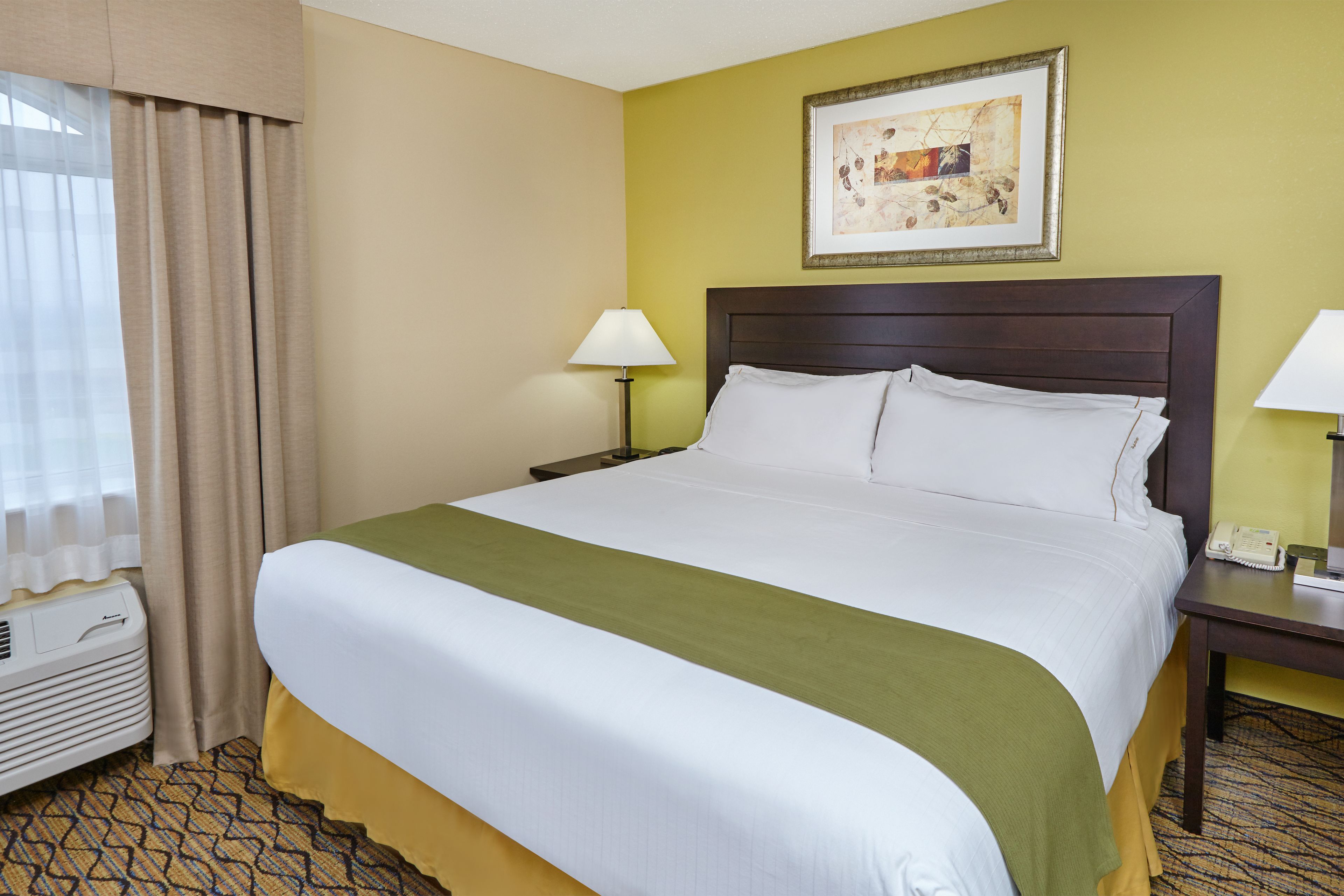 A hotel room with a large bed, a bedside table with a lamp, a framed picture on the wall, and a window with curtains.