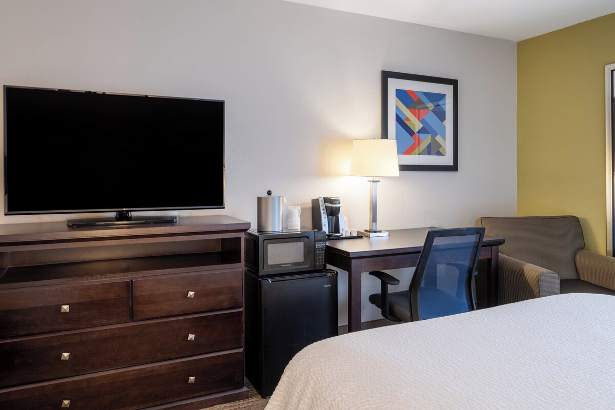 A hotel room with a flat-screen TV, a desk with a lamp, a coffee maker, a microwave, a mini fridge, a bed, and a chair.