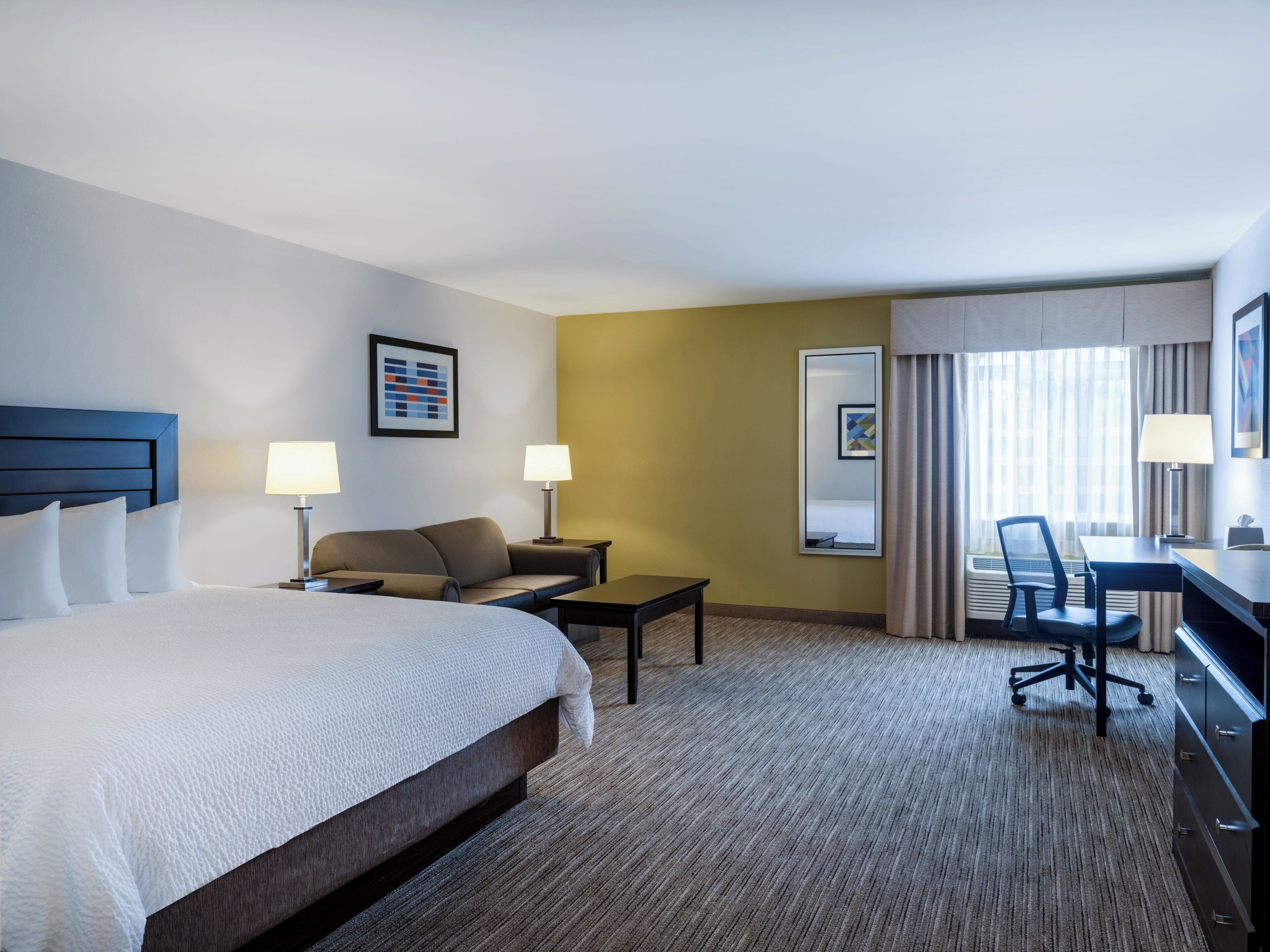 A hotel room with a large bed, two bedside tables with lamps, a sofa, a desk with a chair, and a large mirror.