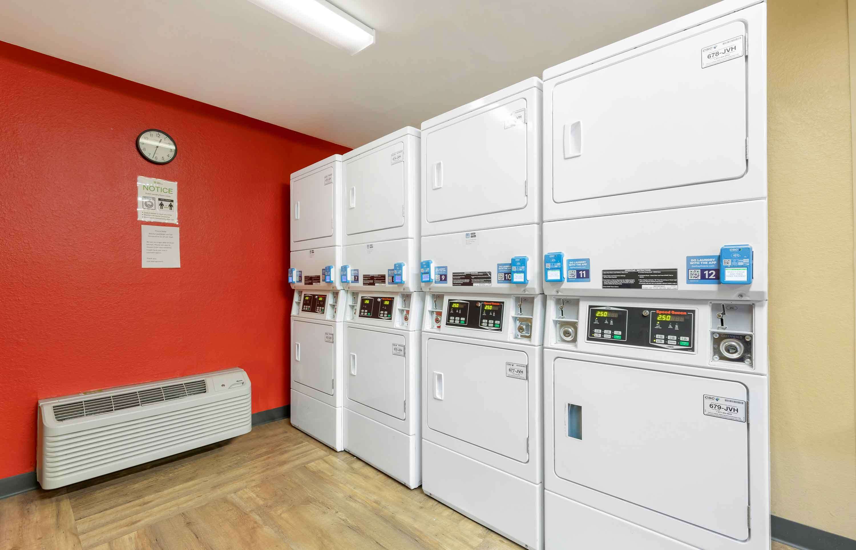 A laundry room with multiple white dryers, a clock, and a notice on the wall.