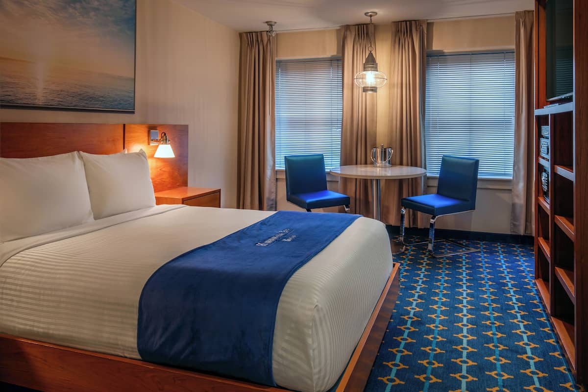 A hotel room with a large bed, a small table with two chairs, a bookshelf, and a painting on the wall.