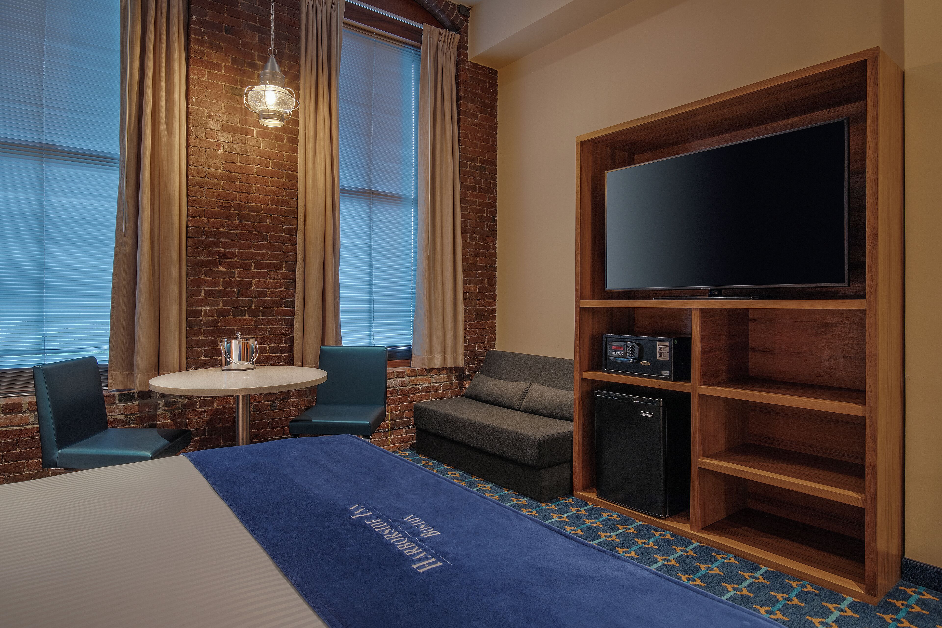 A hotel room with a brick wall, a flat-screen TV, a small table, two chairs, a sofa, and a minibar.