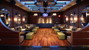 A spacious lounge area with a wooden floor, multiple seating options including sofas and ottomans, a television screen, and a ceiling with circular light fixtures.