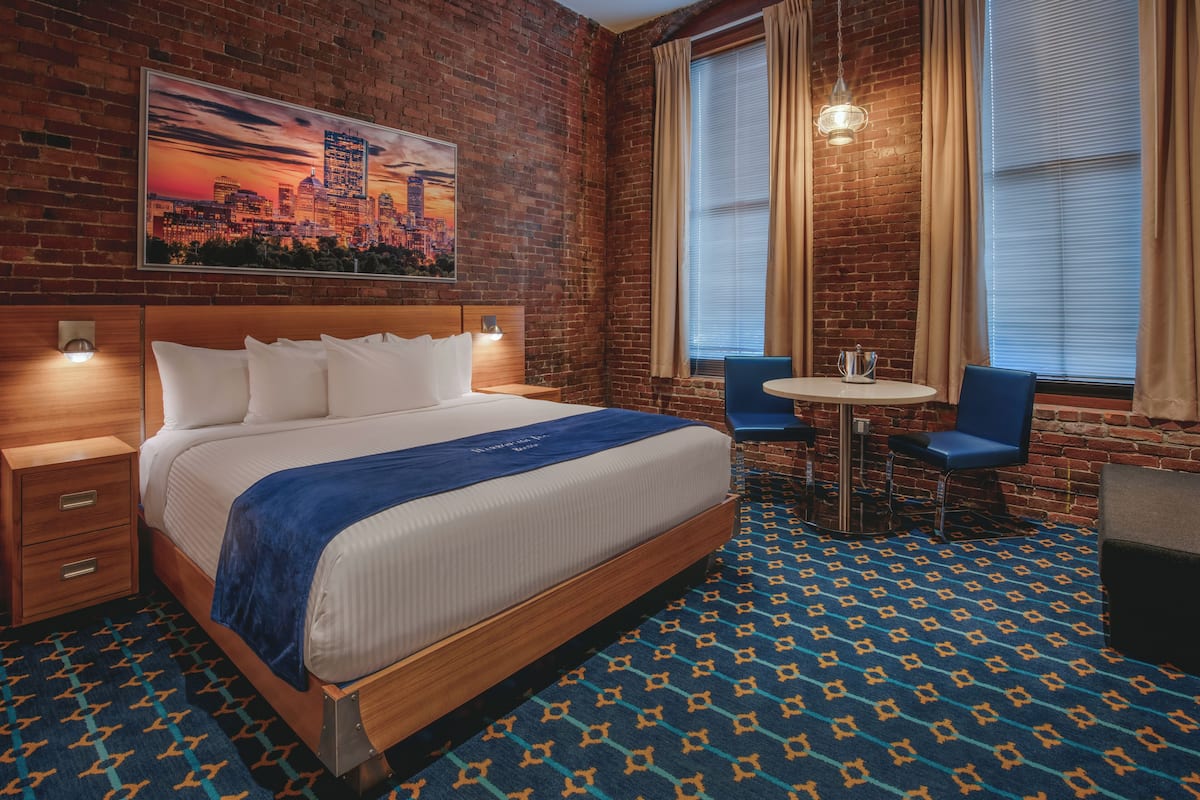 A hotel room with a large bed, a cityscape painting, a small table with chairs, and a patterned carpet.