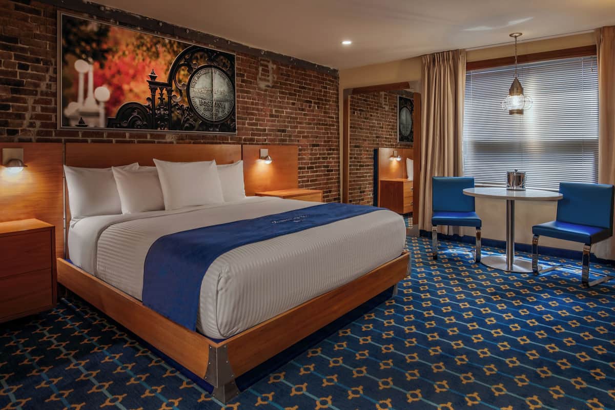 A hotel room with a large bed, a small table, two chairs, a brick wall, and a window with blinds.