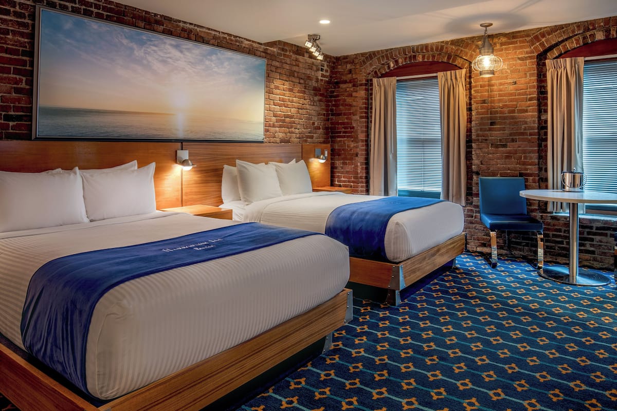 A hotel room with two beds, a large painting of a sunset, a brick wall, a desk with a chair, and a patterned carpet.