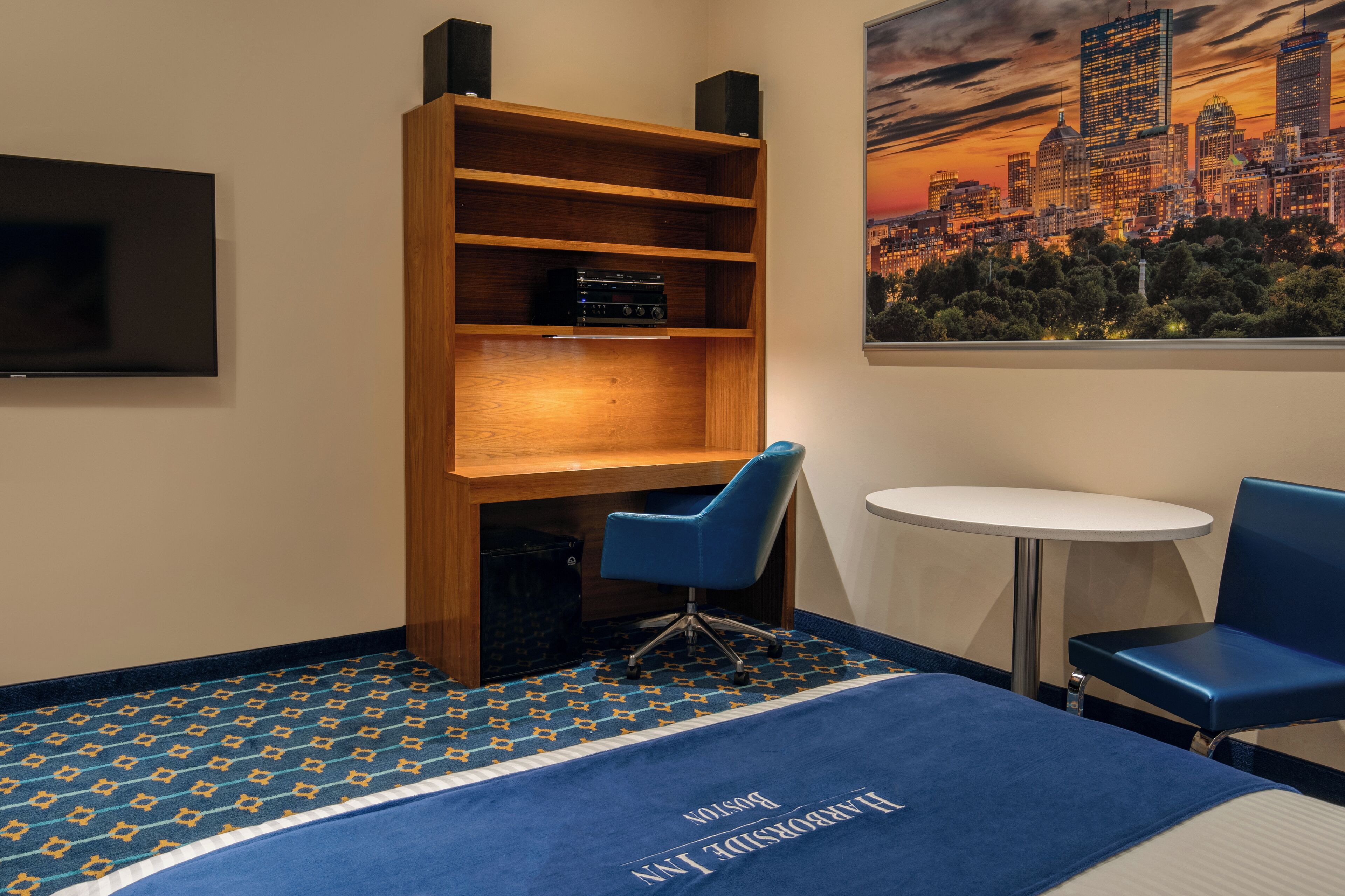 A hotel room with a bed, a bookshelf, a desk, two chairs, a TV, and a cityscape painting.
