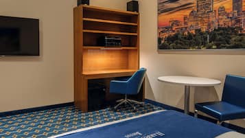 Habitación de hotel con una cama, una estantería, un escritorio, dos sillas, un televisor y una pintura de un paisaje urbano.