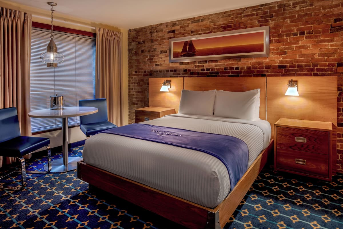 A hotel room with a brick wall, a bed with a blue headboard, two bedside tables with lamps, a small round table with a chair, and a window with curtains.