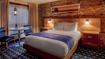 Habitación de hotel con pared de ladrillo, una cama con cabecera azul, dos mesitas de noche con lámparas, una pequeña mesa redonda con una silla y una ventana con cortinas.