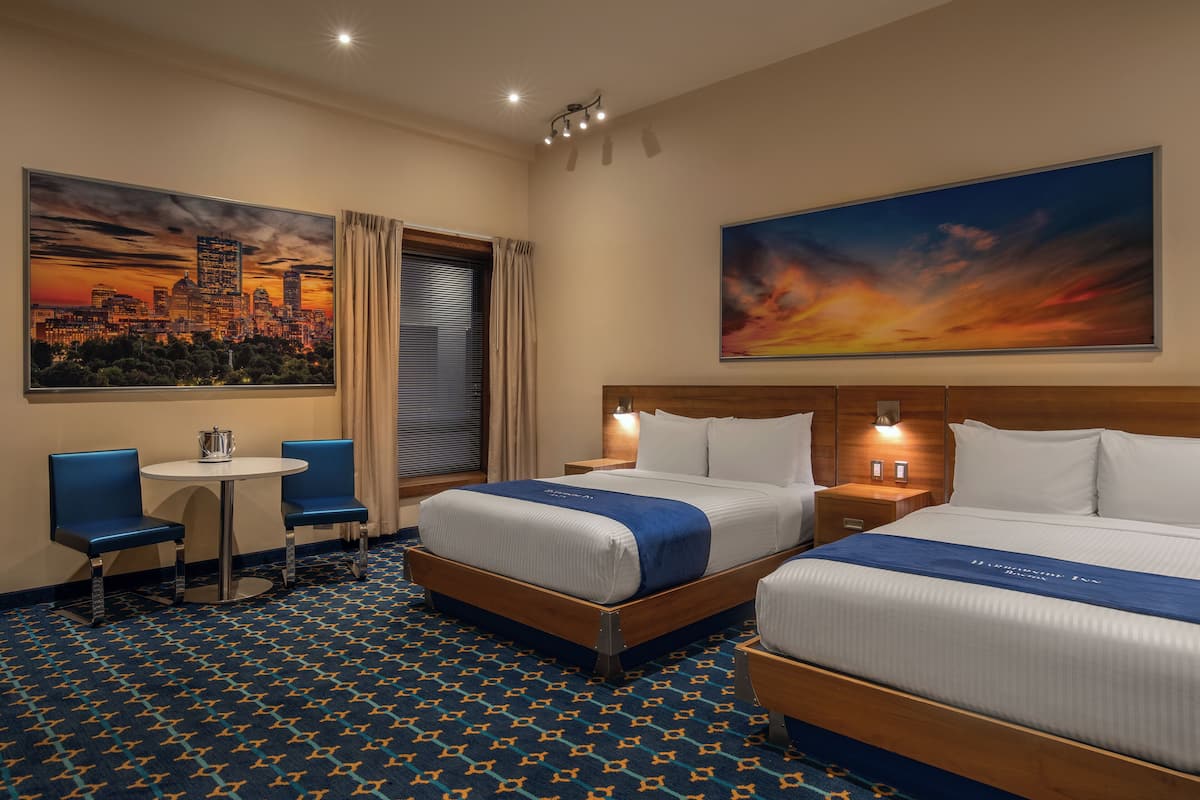 A hotel room with two beds, a small table, two chairs, and a cityscape painting on the wall.