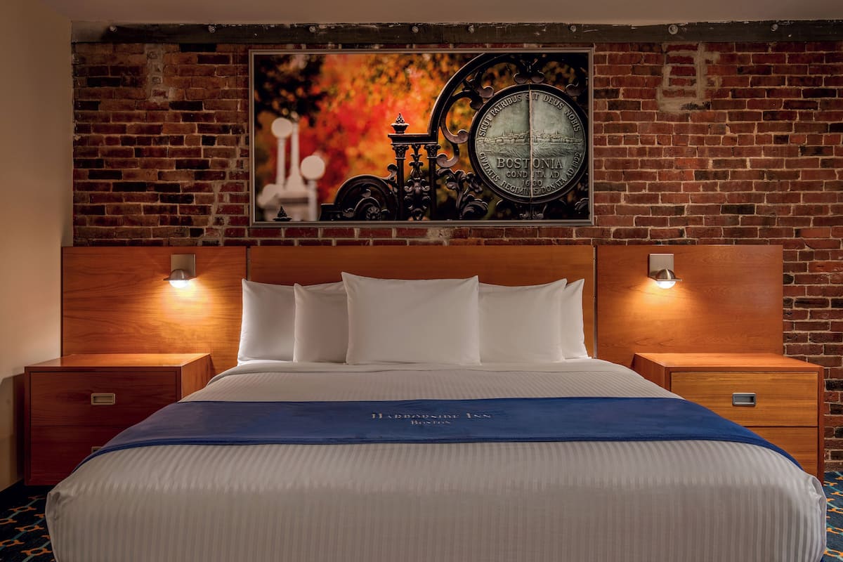 A hotel room with a brick wall, a large bed with white bedding, two bedside tables with lamps, and a framed picture above the bed.